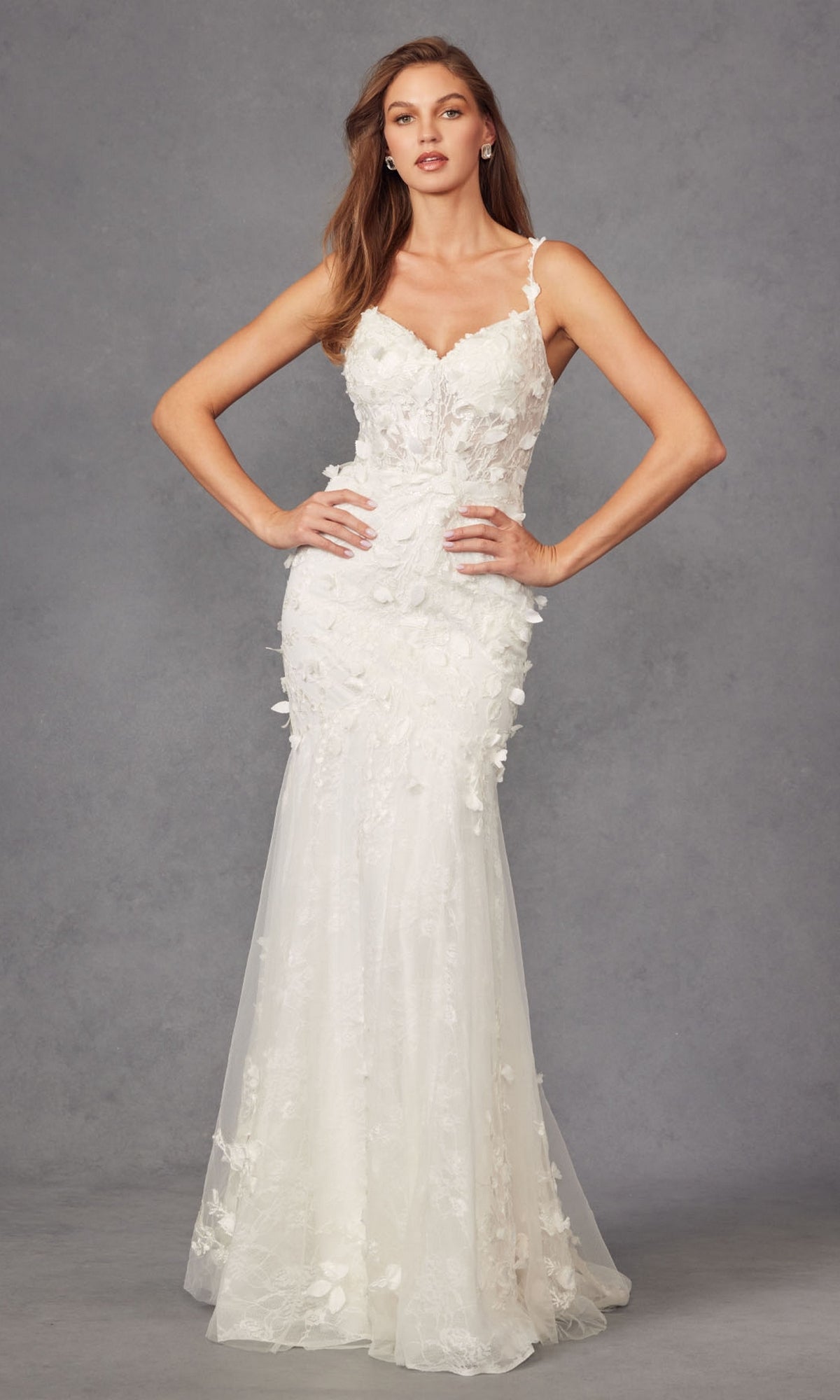 A woman wearing the Long Prom Dress JT2475UW by Juliet, an off-white mermaid gown with delicate floral appliqué, stands against a plain gray background.